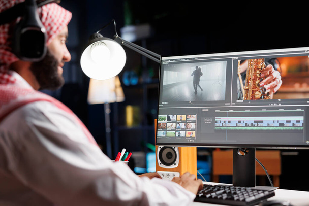 Arabische männliche Filmemacher, die am Tisch sitzen und fleißig an der Postproduktion für einen Film arbeiten. Muslimischer Videoredakteur in traditioneller Kleidung, der kabellose Kopfhörer trägt, während er Filmmaterial auf dem Computerbildschirm betrachtet. - Foto, Bild