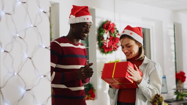Žena v klobouku Santa překvapen kolega s promyšlený vánoční dárek při práci na notebooku ve slavnostní zdobené kanceláři. Kolegyně nabízející pracovnici přítomné a objímající ji během prázdninového období - Fotografie, Obrázek