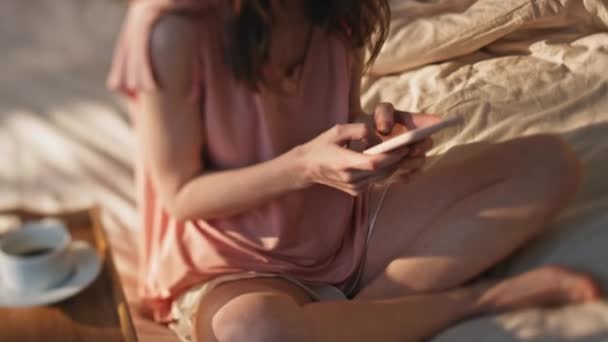 Closeup hands typing smartphone screen. Smiling girl resting in bed messaging online on cozy weekend. Relaxed woman using mobile phone enjoying hotel breakfast in pajamas. Carefree female in sunlight - Footage, Video