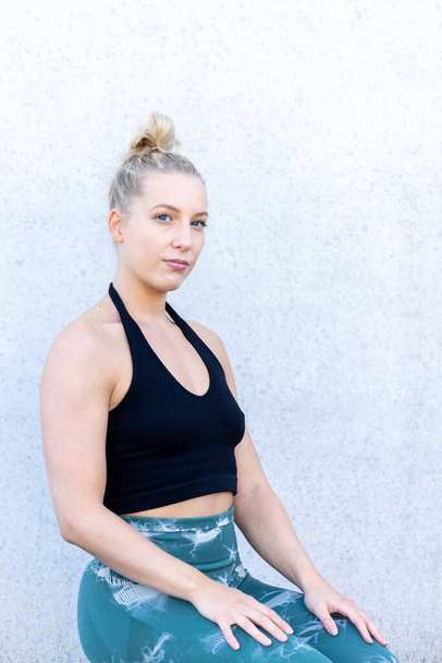 Een jonge blonde blanke vrouw in zwart-blauwe sportkleding zit op haar knieën tegen een betonnen muur en toont vastberaden haar sportieve en actieve levensstijl. Sportieve Millennial vrouw in - Foto, afbeelding