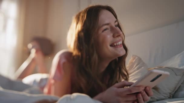 Zrelaksowana dziewczyna trzymająca smartfona leżącego na łóżku. Beztroski śmiech kobiet oglądanie śmieszne treści wideo online za pomocą telefonu komórkowego. Atrakcyjny uśmiechnięty model bawiący się przeglądaniem stron w świetle słonecznym. - Materiał filmowy, wideo