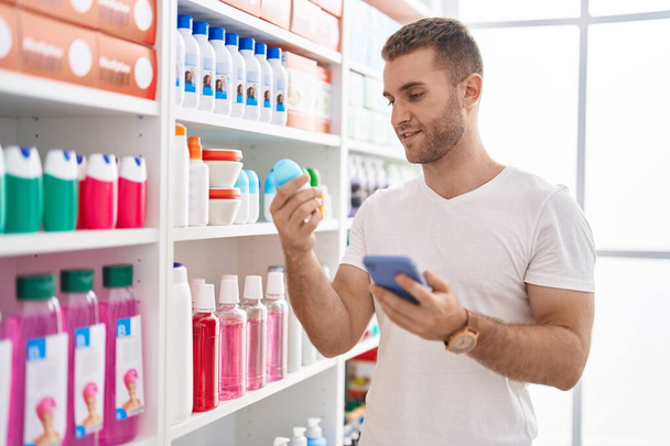 Jovem caucasiano homem cliente usando smartphone segurando desodorizante na farmácia - Foto, Imagem