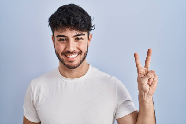 Hispanischer Mann mit Bart steht auf weißem Hintergrund und zeigt mit Fingern Nummer zwei nach oben, während er selbstbewusst und glücklich lächelt.  - Foto, Bild