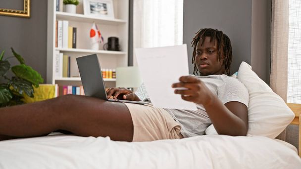 Afro-Américain utilisant un ordinateur portable document de lecture couché sur le lit dans la chambre - Photo, image