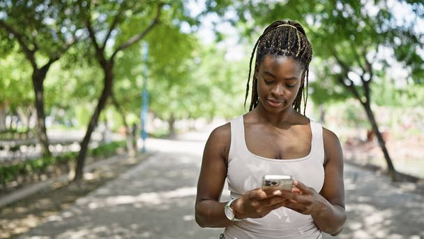 Afroamerykanka używa smartfona z poważnym wyrazem twarzy w parku - Zdjęcie, obraz