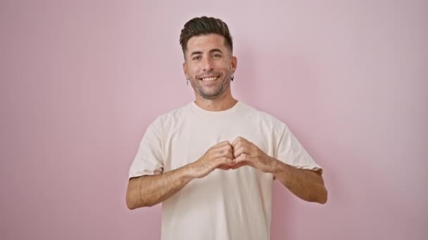 Mosolygó fiatal spanyol férfi teszi magabiztos szív gesztus a kezét az elszigetelt rózsaszín háttér, megjelenítve vidám bizalmat divat-alkalmi életmód - Felvétel, videó