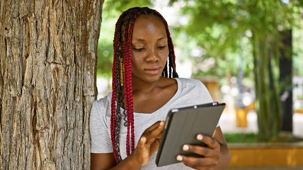 Cool african american woman focused on her touchpad, leaning on tree in city park, casual lifestyle vibe immersed in green outdoors - Photo, Image