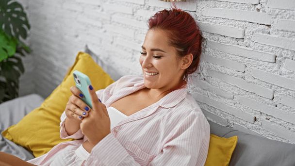 Joven mujer pelirroja hermosa, cómodamente relajada en pijama, sonriendo mientras usa el teléfono inteligente en la cama acogedora, disfrutando de una mañana tranquila en el interior de su dormitorio hogareño - Foto, imagen