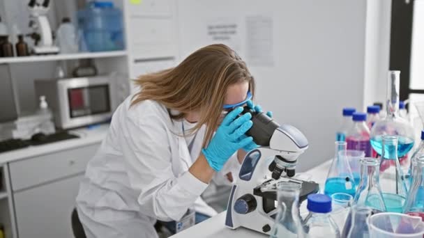 Mujer rubia joven molesta científica lucha con el problema de la investigación, trabajando incansablemente en el análisis utilizando el microscopio en el laboratorio - Imágenes, Vídeo