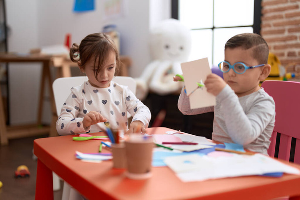 Очаровательная девушка и мальчик сидят на столе и режут бумагу в детском саду - Фото, изображение