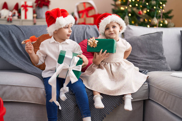 Dos niños sosteniendo regalos sentados en el sofá por el árbol de Navidad en casa - Foto, Imagen