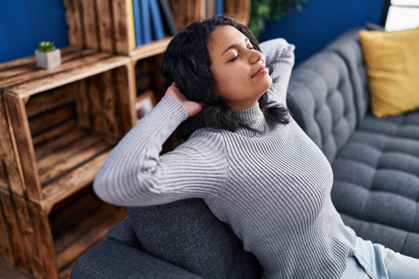 Mujer latina joven relajada con las manos en la cabeza sentada en el sofá en casa - Foto, Imagen