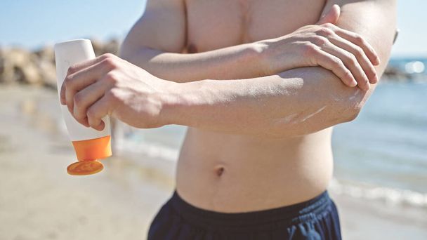 Молодий іспаномовний чоловік турист наносить сонцезахисний крем на руку на узбережжі
 - Фото, зображення