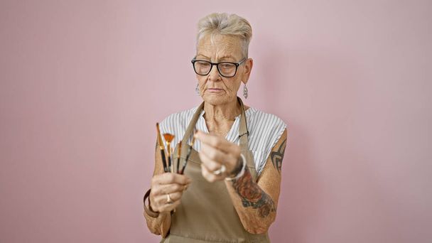 真面目な,灰色の髪のシニア女性アーティストは,ピンクの上にペンキブラシを集中的に保持しています, 孤立した背景 - フルスイングで絵画クラス - 写真・画像
