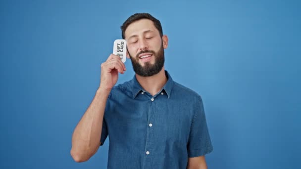 Jonge Spaanse man glimlachend zelfverzekerd wijzend naar gift card doen duim omhoog gebaar over geïsoleerde blauwe achtergrond - Video