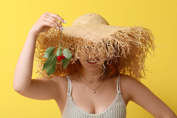 Šťastná krásná mladá žena v proutěném klobouku s větev zralé třešně na žlutém pozadí - Fotografie, Obrázek