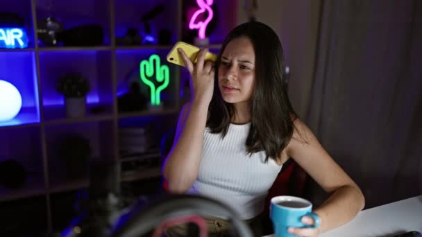 ストリームの夜 - 若くて美しいヒスパニック系女性ストリーマーがゲームに魅了され,コーヒーを飲み,ネオンリッターゲームルーム内のスマートフォンで音声メッセージを聞く - 映像、動画