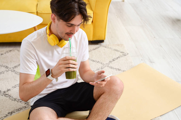 Sportieve jongeman met gezonde smoothie en mobiele telefoon thuis - Foto, afbeelding