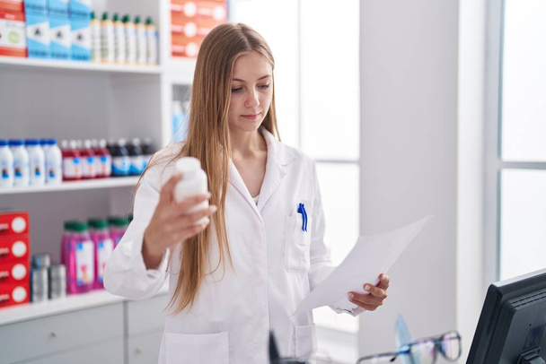 Молода біла жінка фармацевт тримає пляшку таблеток, читаючи рецепт в аптеці
 - Фото, зображення