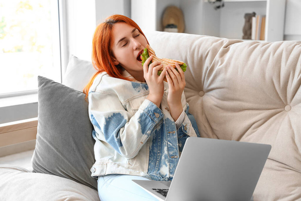 Young redhead woman with laptop eating tasty sandwich at home - Photo, Image