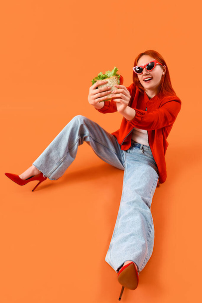 Junge rothaarige Frau mit leckerem Sandwich sitzt auf orangefarbenem Hintergrund - Foto, Bild