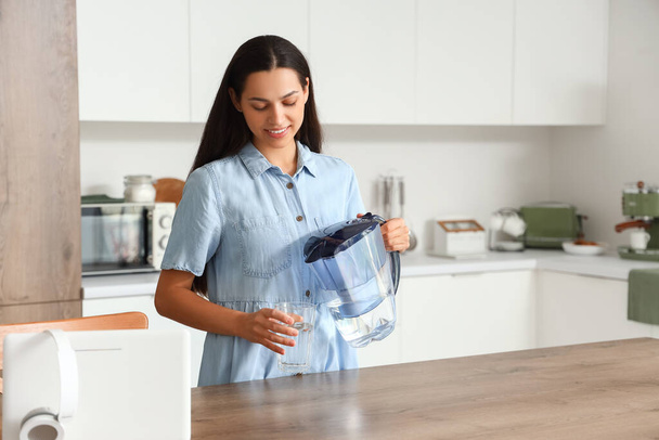 Beautiful young woman pouring pure water from filter jug into glass in kitchen - Photo, Image