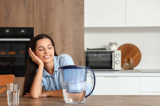 Beautiful young woman with water filter jug and glass in kitchen - Photo, Image