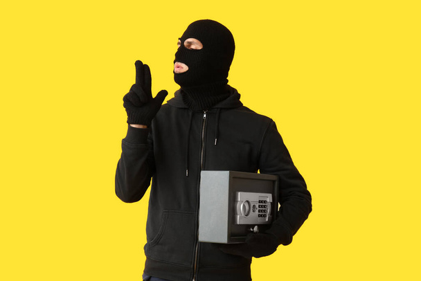 Ladrón masculino con caja fuerte mostrando gesto de pistola sobre fondo amarillo - Foto, imagen