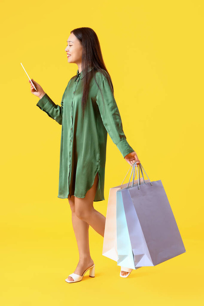 Jeune femme asiatique avec tablette et sacs à provisions sur fond jaune - Photo, image