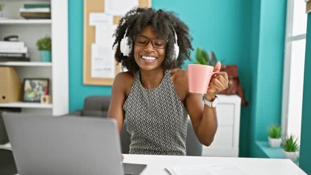 Afrikkalainen amerikkalainen nainen liiketoiminnan työntekijä kuuntelee musiikkia ja juo kahvia toimistossa  - Materiaali, video