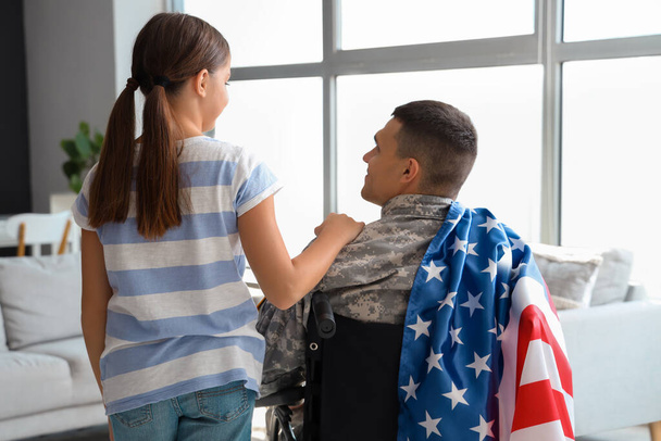 Солдат в инвалидном кресле со своей маленькой дочерью дома, вид сзади. Празднование Дня ветеранов - Фото, изображение