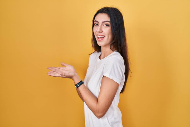 Mladá krásná žena stojící nad žlutým pozadím ukazuje stranou s rukama otevřenýma dlaněmi ukazující kopírovací prostor, prezentující reklamu s nadšeným úsměvem  - Fotografie, Obrázek