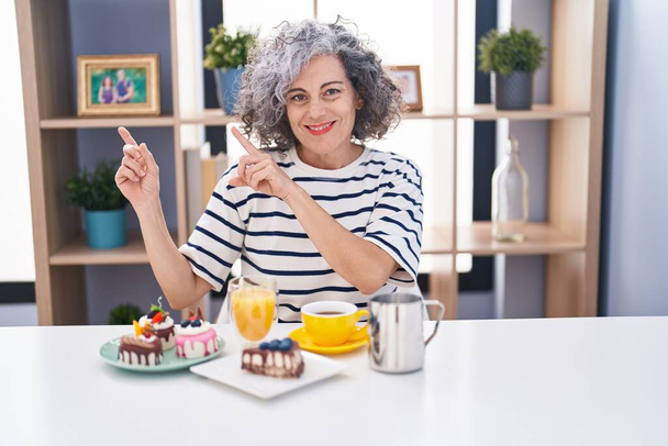 Frau mittleren Alters mit grauen Haaren isst Gebäck und trinkt Kaffee zum Frühstück, lächelt und blickt in die Kamera, die mit zwei Händen und Fingern zur Seite zeigt..  - Foto, Bild