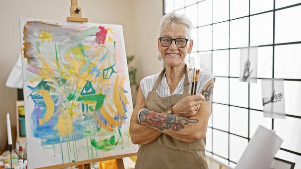 Usmívající se šedovlasá starší umělkyně, stojící s uvolněným gestem zkřížených paží, užívající si svého koníčka uprostřed plátna a štětců ve svém ateliéru. - Fotografie, Obrázek