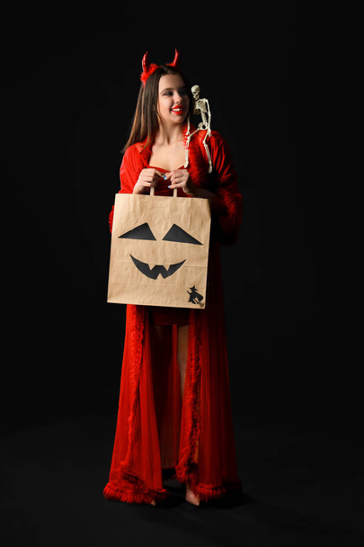 Молода жінка, одягнена на Хеллоуїн як диявол з подарунковою сумкою та скелетом на темному тлі - Фото, зображення