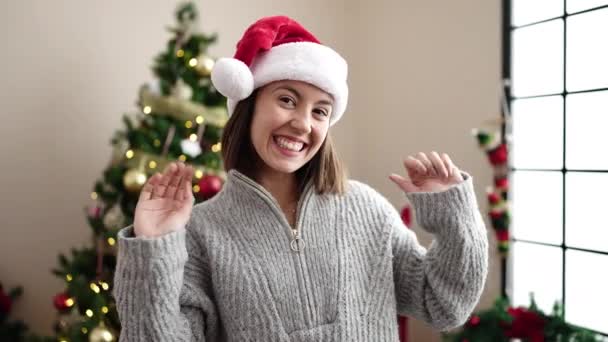 Νεαρή όμορφη ισπανόφωνη γυναίκα χαμογελά με αυτοπεποίθηση στέκεται δίπλα στο χριστουγεννιάτικο δέντρο στο σπίτι - Πλάνα, βίντεο