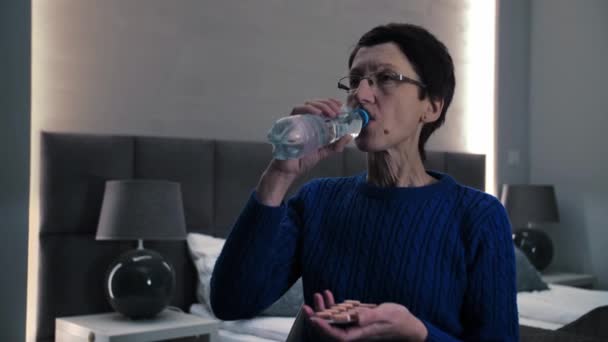 Starší žena si vzít pilulky sedí na gauči. Dospělá dáma drží léky pilulky a pitnou vodu z láhve v temné ložnici interiéru. Vysoce kvalitní 4K záběry - Záběry, video