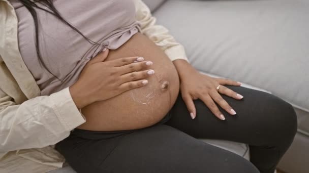 Nenucená, bezstarostná mladá těhotná žena radostně aplikuje ošetření kůže na břicho, zatímco sedí doma na pohovce a těší se ze svého mateřství - Záběry, video
