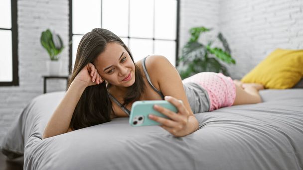 Wesoła młoda latynoska kobieta leżąca w łóżku, w piżamie, mając szczęśliwy telefon wideo w przytulnej sypialni swojego mieszkania - Zdjęcie, obraz