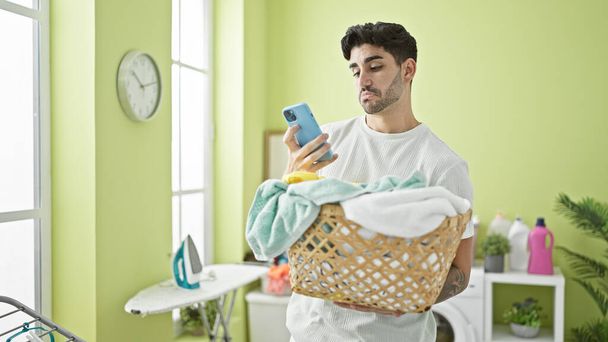 Jonge Spaanse man houdt mand met kleding met behulp van smartphone in de wasruimte - Foto, afbeelding