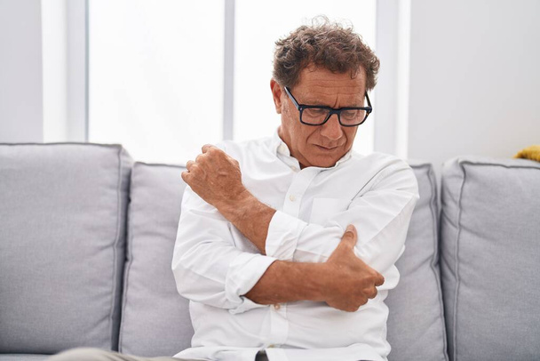 Uomo di mezza età che soffre di dolore al gomito seduto sul divano a casa - Foto, immagini