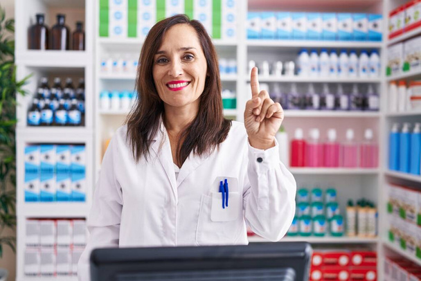 Middelbare leeftijd brunette vrouw werken bij apotheek drogisterij tonen en wijzen met vinger nummer een, terwijl glimlachen zelfverzekerd en gelukkig.  - Foto, afbeelding