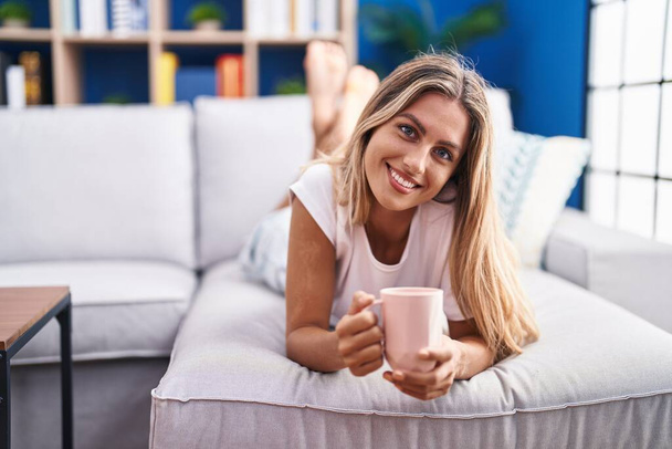 Nuori blondi nainen juo kahvia makaa sohvalla kotona - Valokuva, kuva
