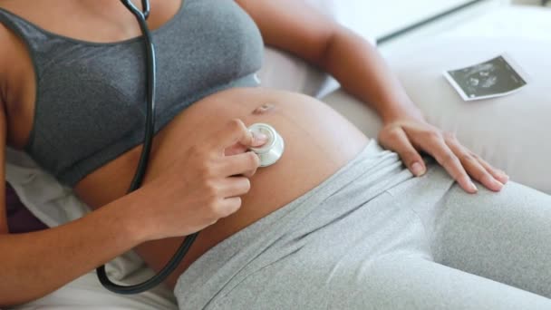 La madre thailandese-afroamericana incinta utilizza felicemente lo stetoscopio medico, ascolta il battito cardiaco del suo feto e posiziona eccitatamente le immagini ecografiche sul letto mentre il bambino si muove nell'utero.. - Filmati, video