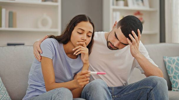Schönes Paar, das sich umarmt, zu Hause verärgert ist, einen positiven Schwangerschaftstest in der Hand hält, ein unerwartetes Familienproblem in ihrem gemütlichen Wohnzimmer - Foto, Bild