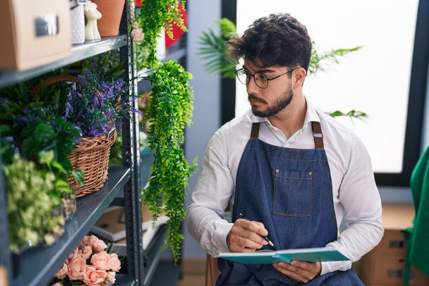 Joven florista hispano escribiendo en cuaderno mirando estanterías en floristería - Foto, imagen