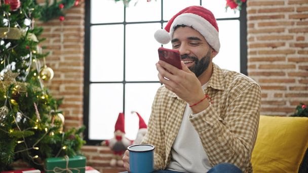 Young hispanic man sending voice message by smartphone drinking coffee celebrating christmas at home - Photo, Image