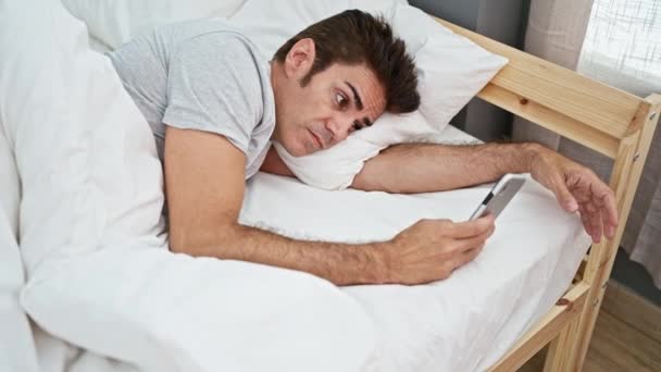 Νεαρός Ισπανός ξαπλωμένος στο κρεβάτι με λυπητερή έκφραση χρησιμοποιώντας smartphone στο υπνοδωμάτιο - Πλάνα, βίντεο
