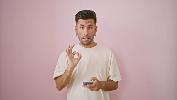 Junger, attraktiver hispanischer Typ, lässig vor rosafarbenem Hintergrund stehend, die "ok" -Handgeste schaukelnd, darin vertieft, eine Nachricht auf sein Smartphone zu tippen, total ernst, aber positiv wirkend. - Foto, Bild