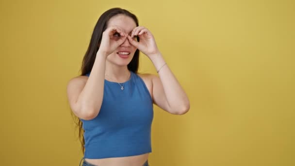 Νεαρή όμορφη ισπανόφωνη γυναίκα χαμογελά με αυτοπεποίθηση κάνοντας κίνηση με κιάλια πάνω από απομονωμένο κίτρινο φόντο - Πλάνα, βίντεο